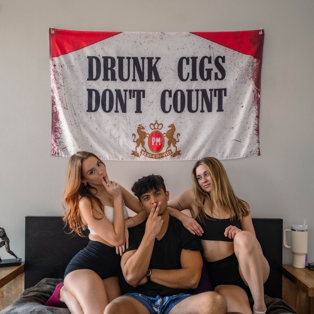 Three friends posing in front of a red and white novelty flag with playful text, in a dorm room setting.