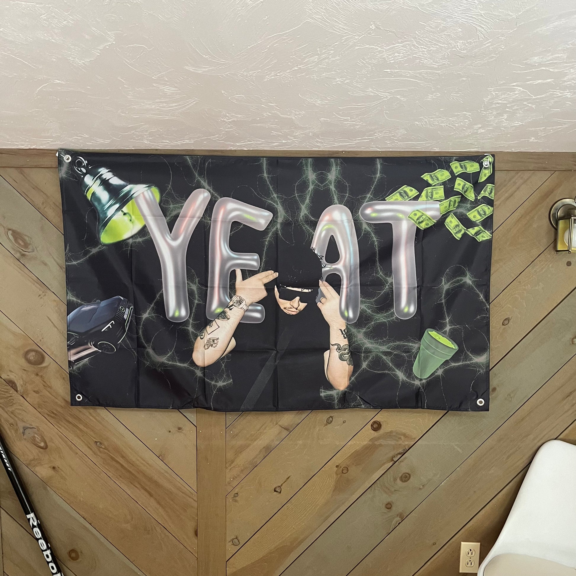 Black flag featuring Yeat in the center with metallic balloon-style letters spelling 'YEAT,' accented by images of cash, a car, a green cup, and an overhead lamp. The background has a neon lightning effect, adding a vibrant and dynamic feel, perfect for fans of the rapper